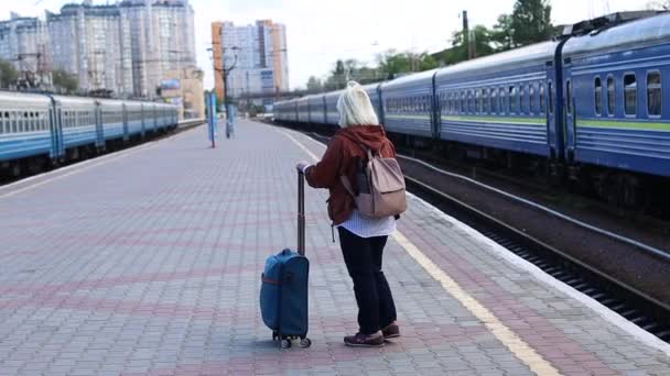 Mujer rubia adulta viajera de pie con equipaje en la estación de tren. Concepto de viaje - Metraje, vídeo