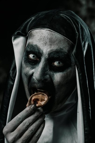 primer plano de una monja malvada asustadiza comiendo un dedo humano con sus dientes ensangrentados, usando un hábito típico blanco y negro - Foto, imagen