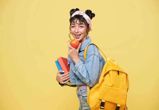 Happy Asian girl holding apple and notebook over yellow background. back to school concept. - Photo, Image