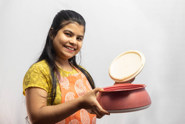 Una bonita ama de casa india en delantal de cocina con cazuela de servicio de apertura sobre fondo blanco - Foto, Imagen