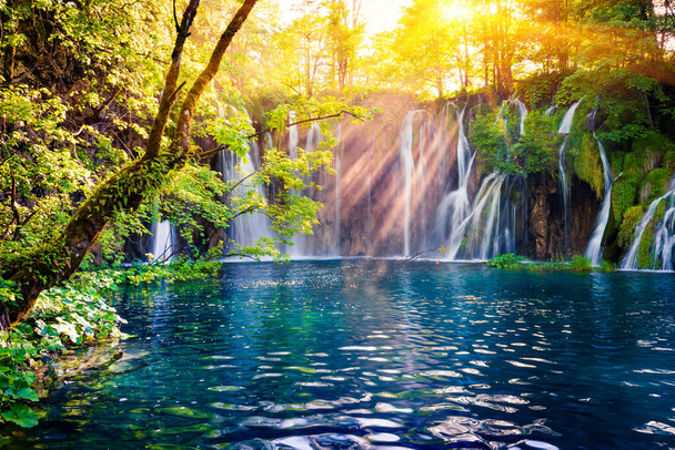 Ostatnie światło słoneczne oświetla wodospad czystej wody w Parku Narodowym Plitwice. Kolorowa wiosenna scena zielonego lasu z błękitnym jeziorem. Wspaniały widok na okolicę Chorwacji, Europy. Piękno natury koncepcja tła. - Zdjęcie, obraz