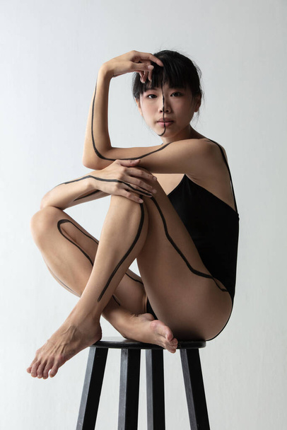 Portrait of young adorable Japanese ballerina sitting on high chair and thinking isolated on light gray studio background. Beauty, line art, inner world concept. - Fotó, kép