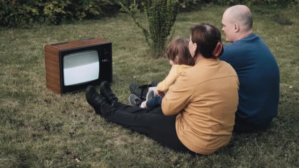 Family with small child are sitting on the grass and watching an old retro TV - Footage, Video
