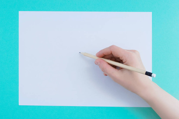 Biała kartka papieru i drewniany ołówek w dłoni białej kobiety na turkusowym tle. - Zdjęcie, obraz