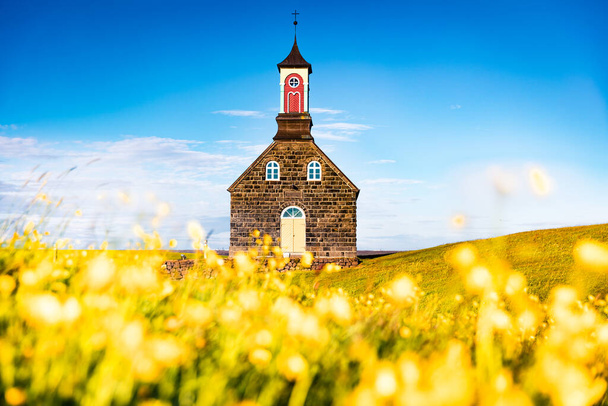 Ηλιόλουστη θέα το πρωί της εκκλησίας Hvalsneskirkja μεταξύ ανθισμένα κίτρινα λουλούδια. Εκπληκτική καλοκαιρινή σκηνή της Ισλανδίας, τοποθεσία Keflavk. Ταξιδεύοντας φόντο έννοια. Ταξίδι στην Ισλανδία. - Φωτογραφία, εικόνα