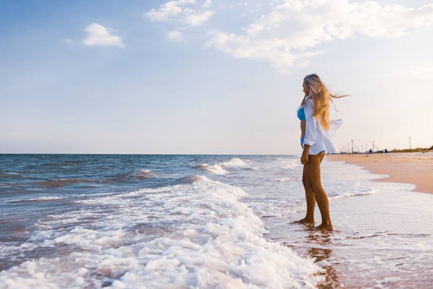 A slender girl in a gentle blue swimsuit and shirt walks along the sandy beach near the blue sea - Fotografie, Obrázek