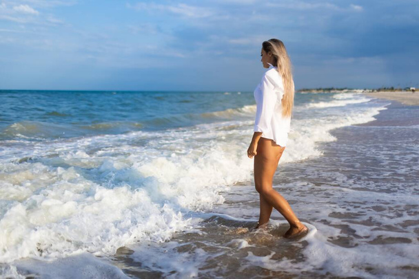 A slender girl in a gentle blue swimsuit and shirt walks along the sandy beach near the blue sea - Foto, afbeelding