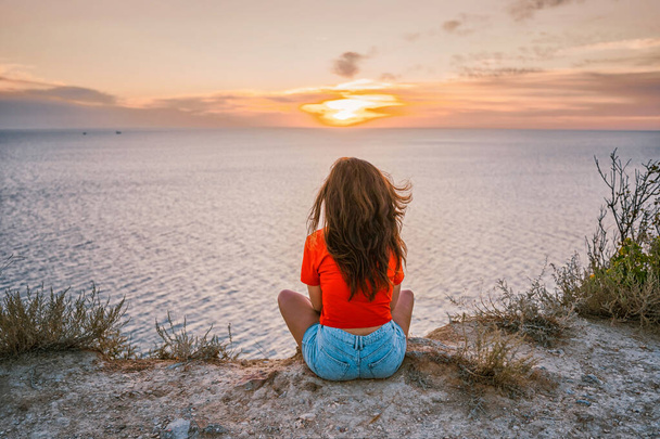  Arkaya bakan kırmızı gömlekli genç bir kadın denizin önündeki uçurumun kenarında oturuyor. Gün batımının güzelliği ile. - Fotoğraf, Görsel