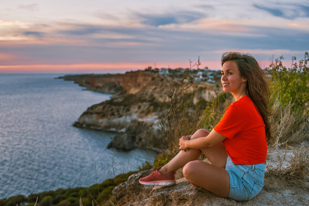 Młoda kobieta turysta stoi na brzegu wysoko nad morzem o zachodzie słońca i podziwia Cape Fiolent na Krymie - Zdjęcie, obraz