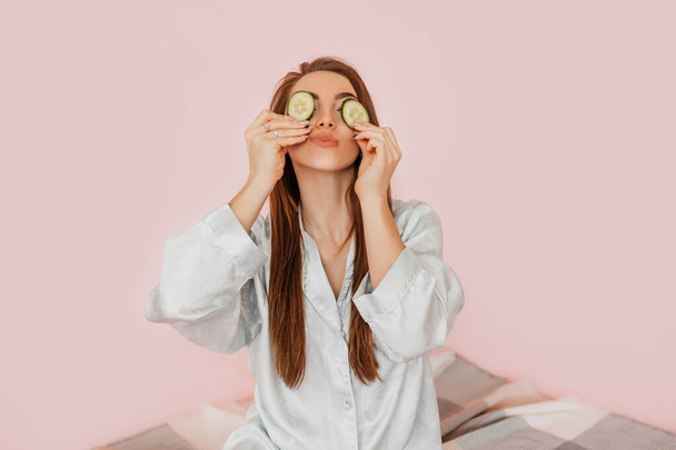 Girl make homemade face beauty masks. Cucumbers for the freshness of the skin around the eyes. Woman take care of youthful skin. Model laughing and having fun in spa on pink background - Фото, зображення