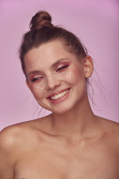Fashion creative make up. Young woman face expression with clean fresh skin and bright make up with rhinestones freckles on her face posing on a bright pink background - Fotografie, Obrázek