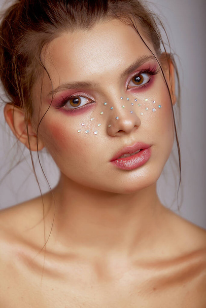 Mode kreativ schminken. Schöne Porträt einer jungen Frau mit sauberer, frischer Haut und hellem Make-up mit Strass Sommersprossen auf ihrem Gesicht posiert auf einem grauen Hintergrund - Foto, Bild