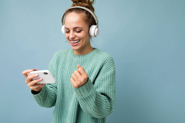 Side-profile photo of attractive emotional positive young woman wearing blue sweater isolated over blue background wearing white bluetooth wireless headphones and listening to music and using mobile - Photo, Image