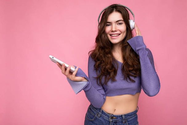 Фото красивой веселой улыбчивой молодой женщины в стильной повседневной одежде на фоне стены, держащей и использующей мобильный телефон в белых наушниках Bluetooth, слушающей музыку и - Фото, изображение