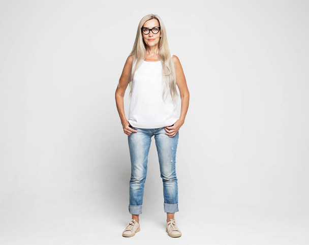Full length photo of stylish elderly woman wearing jeans, white blouse and glasses - 写真・画像