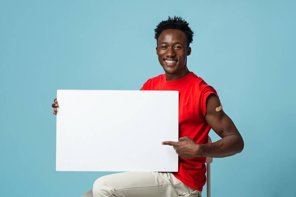 Información de vacunación. Sonriente chico afroamericano con yeso en el brazo sosteniendo la placa en blanco - Foto, imagen