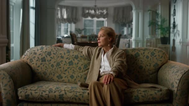 Femme âgée réfléchie assise sur le canapé dans une maison de luxe. Femme mûre pensée  - Séquence, vidéo