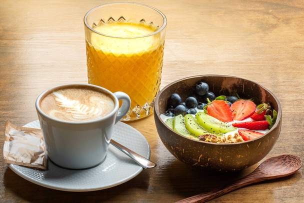 Primo piano di sana colazione con ciotola di frutta fresca a fette e yogurt biologico. Succo d'arancia fresco naturale e caffè cremoso con schiuma sul tavolo di legno. - Foto, immagini