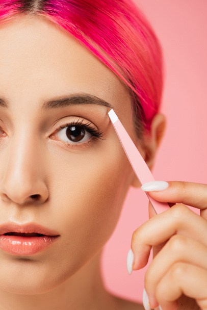close up of young woman with dyed hair holding tweezers while shaping eyebrow isolated on pink  - Photo, Image