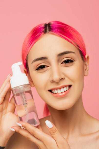 tattooed young woman with colorful hair smiling while holding bottle with cleansing product isolated on pink  - Foto, afbeelding