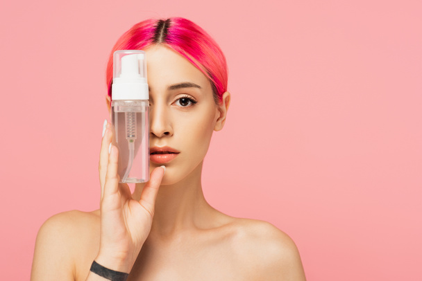 wytatuowana młoda kobieta z kolorowymi włosami trzymająca butelkę z produktem oczyszczającym, zakrywająca oko odizolowane na różowo  - Zdjęcie, obraz