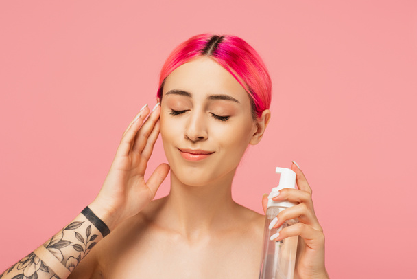 getatoeëerde jonge vrouw met geverfd haar en gesloten ogen houden fles met reinigingsmiddel terwijl glimlachen geïsoleerd op roze  - Foto, afbeelding