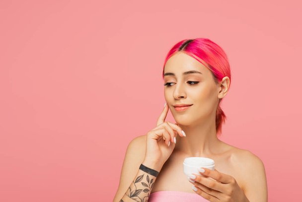 tattooed young woman with colorful hair smiling while holding container and applying face cream isolated on pink - Foto, imagen