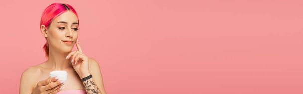 tetovált fiatal nő színes haj mosolyog, miközben gazdaság tartály és alkalmazása arckrém izolált rózsaszín, banner - Fotó, kép