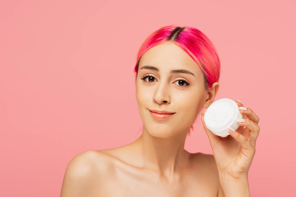 positive young woman with colorful hair smiling while holding container with face cream isolated on pink - 写真・画像