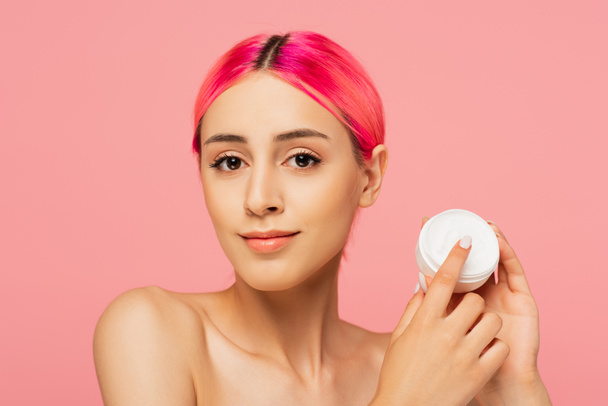 joyful young woman with dyed hair holding container with cosmetic cream isolated on pink  - Foto, Bild