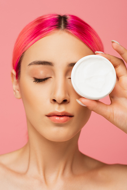 young woman with colorful hair covering eye while holding container with cosmetic cream isolated on pink - Foto, Bild