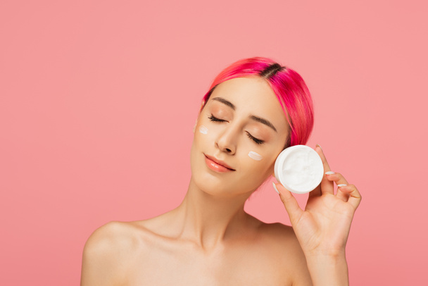 pleased young woman with colorful hair and cream on cheeks holding container isolated on pink - 写真・画像