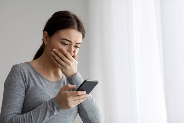 Aufgebracht depressive Millennial hübsche Frau bedeckt ihren Mund mit der Hand und schaut auf Telefon - Foto, Bild