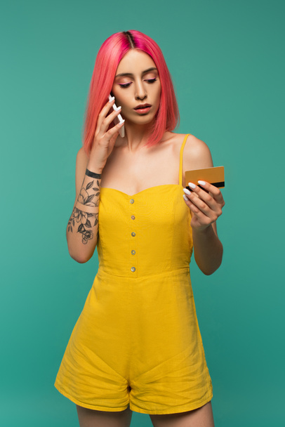 jeune femme tatouée avec des cheveux teints en rose tenant la carte de crédit et ayant un appel téléphonique isolé sur turquoise - Photo, image