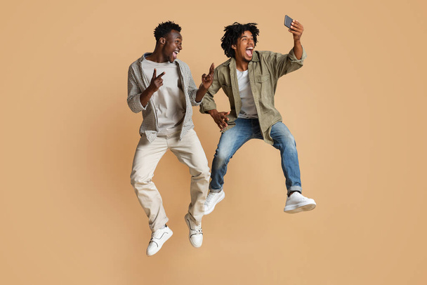 Samen voor de gek houden. Twee zwarte jongens nemen selfie terwijl springen over Beige achtergrond - Foto, afbeelding