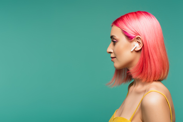 vue latérale de la jeune femme avec des cheveux teints en rose dans un écouteur sans fil isolé sur bleu - Photo, image