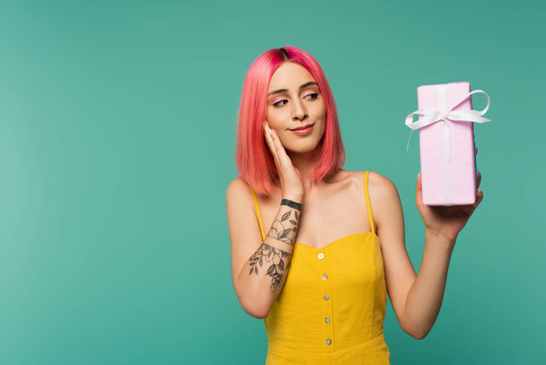 довольная молодая женщина с розовыми окрашенными волосами, держащих завернутый подарок изолирован на синий  - Фото, изображение