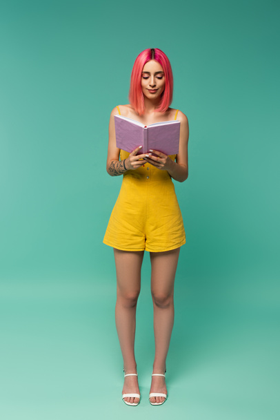 full length of young woman with pink dyed hair reading book on blue  - Zdjęcie, obraz