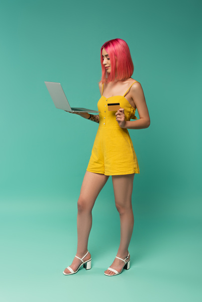 pełna długość młodej kobiety z różowymi farbowanymi włosami trzymającej kartę kredytową i laptopa na niebiesko  - Zdjęcie, obraz