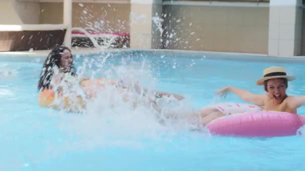 Womens friendship and fun entertainment. Two funny energetic girlfriends relaxing in pool of luxury hotel, swimming on large inflatable lifebuoys, laughing splashing water, swinging legs waving hands - Footage, Video