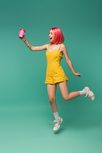 full length of smiling young woman with pink dyed hair jumping with paper cup on blue  - Foto, immagini