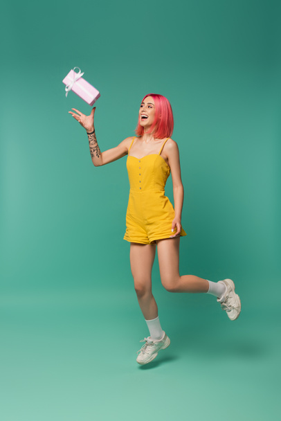 full length of smiling young woman with pink dyed hair jumping and throwing present on blue  - Fotografie, Obrázek