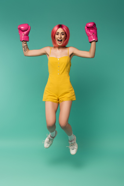 full length of amazed young woman with pink dyed hair in boxing gloves jumping on blue  - Φωτογραφία, εικόνα