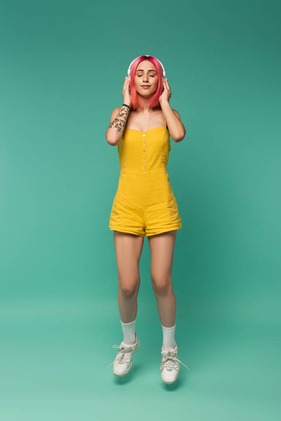 full length of young woman with pink dyed hair and wireless headphones levitating on blue  - Photo, Image