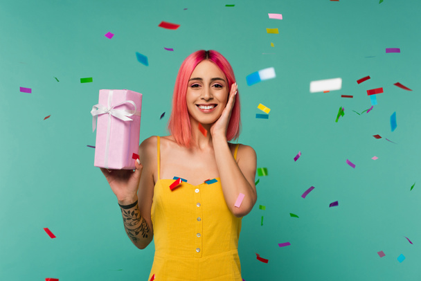 счастливая молодая женщина с розовыми окрашенными волосами держа подарочную коробку рядом красочные конфетти на голубом - Фото, изображение