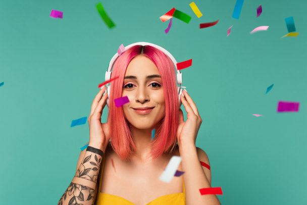 щаслива молода жінка з рожевим фарбованим волоссям в бездротових навушниках біля барвистих конфетті на синьому
 - Фото, зображення