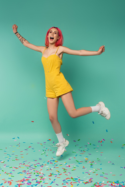full length of positive young woman with pink dyed hair jumping near colorful confetti on blue  - Foto, immagini