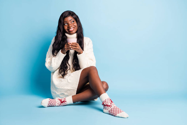 Photo of adorable dark skin lady wear white winter outfit sitting floor coffee looking empty space isolated blue color background - Zdjęcie, obraz