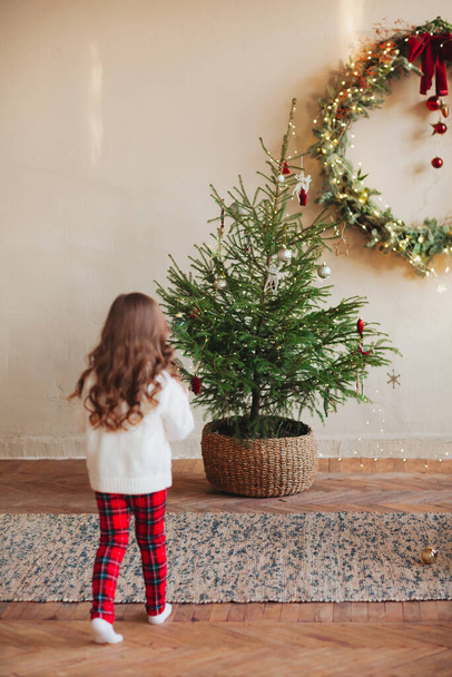 Cute little girl on carpet under by decorated christmas tree during winter holiday season, wearing white buttoned sweater with plaid pants - Foto, Bild