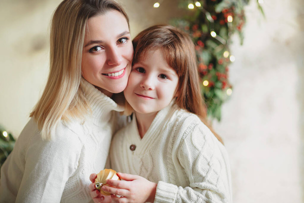 La madre allegra e la figlia di ragazza bella in maglioni lavorati a maglia accoglienti che decorano l'abete di Natale con bauble di anno nuovo e giocattoli in soggiorno, sorridono l'atmosfera di sentimento di vacanza invernale. Vacanze in famiglia - Foto, immagini